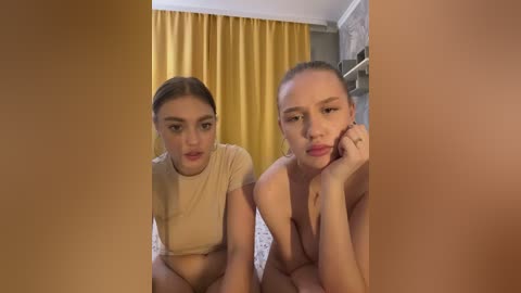 Video of two young women, both with light skin and straight, light brown hair, wearing beige crop tops. They are looking forward, with one resting her chin on her hand, the other appearing pensive. Background features yellow curtains and a bed.