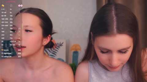 Media: Video of two young women with pale skin, one with dark hair in a ponytail, the other with straight, light brown hair, both in tank tops. Background shows a blurred, indoor setting with artwork.
