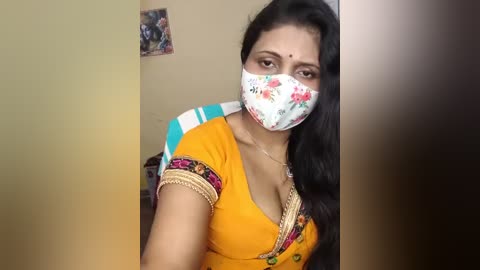 Media: Video of a South Asian woman with medium-brown skin, long black hair, wearing a floral face mask, orange saree with gold trim, and seated indoors against a beige wall.