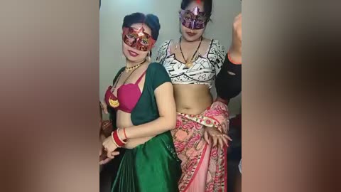 Video of two women in traditional Indian attire, wearing elaborate masks; one in a green sari with a red heart-shaped bra, the other in a white and pink sari with a black blouse.