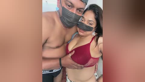 Video of a shirtless man with a mask and a woman in a red bra, both wearing surgical masks, in a close, intimate embrace.