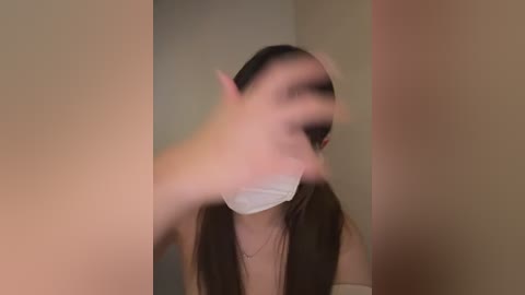 Video of a woman with long dark hair, wearing a white surgical mask, blurred face, and a beige top, captured from a side angle in a dimly lit room.