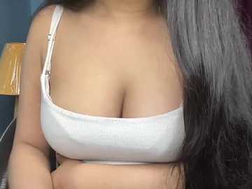 Video of a light-skinned woman with long, dark hair, wearing a white ribbed tank top that accentuates her large breasts. Background shows a wooden chair and part of a wall.