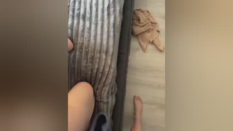 Video of a person in a bed, wearing a beige robe, with a gray, textured blanket, beige towel, and bare feet on a light wooden floor.
