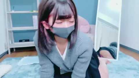 Video of an Asian woman with straight, shoulder-length black hair, wearing a gray cardigan, white blouse, and black skirt. She's kneeling on a light blue rug, gagged with gray tape, in a room with white shelves and a mirror.