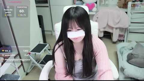 Video of an Asian woman with long black hair, wearing a pink cardigan, sitting on a white chair, with a white cloth over her face, in a cluttered room.