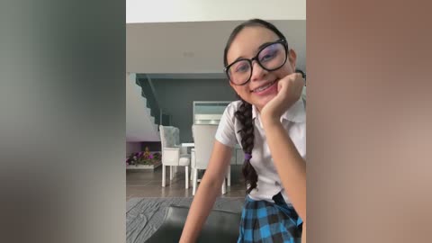 Video of a smiling, dark-haired girl in a white blouse and blue plaid skirt, wearing glasses, in a modern living room with white furniture and green walls.