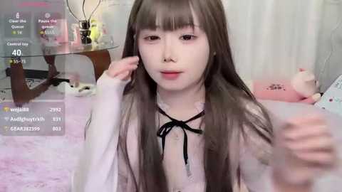 Video of a young Asian woman with long, straight brown hair, wearing a white blouse and black ribbon, sitting on a soft, pink carpet, surrounded by plush toys and a glass table.