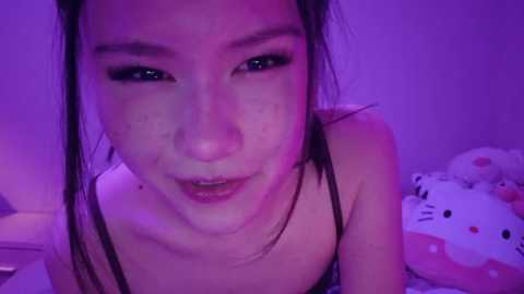 Media: Video of an Asian woman with fair skin and dark hair, wearing a black spaghetti strap top, smiling under purple lighting, surrounded by plush toys.