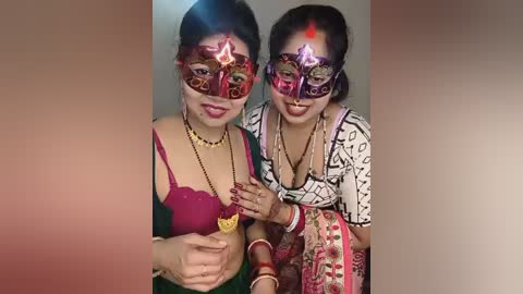 Video of two women in vibrant Indian traditional attire, wearing elaborate red and purple masks, with gold jewelry, in front of a blurred background.