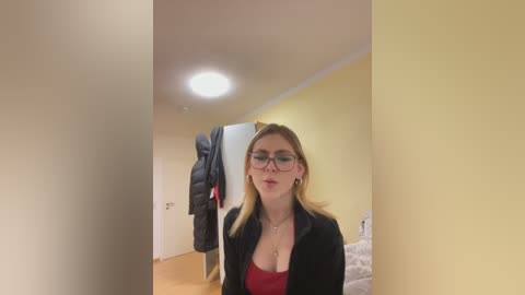 A video of a young Caucasian woman with long blonde hair, wearing glasses, a red top, and a black blazer, standing in a narrow hallway with yellow walls, a hanging jacket, and a visible light bulb.