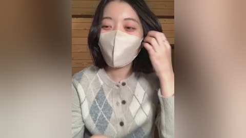 A video of an East Asian woman with shoulder-length black hair, wearing a beige face mask and a gray, diamond-patterned cardigan, against a wooden background.