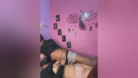 Video of a young woman with medium skin tone, wearing a black mask, lying on a bed with a man performing oral sex on her. The room has pink walls adorned with fairy-themed decorations.