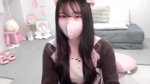 Media: A video of a young woman with long black hair and a pink face mask, dressed in a pink and brown maid outfit, kneeling on a pink cushion in a light-filled, girly room with pink accents.