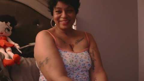 Video of a smiling, tattooed woman with short curly black hair, wearing a colorful tank top, standing in a dimly lit room with a Minnie Mouse plush on a bed.