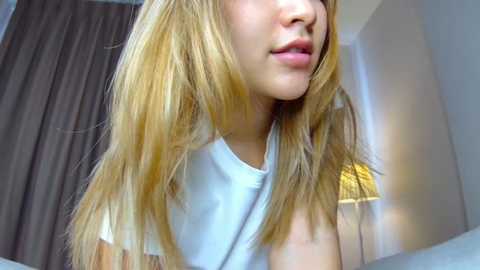 Video of a young woman with long blonde hair, wearing a white t-shirt, looking to the right in a dimly lit room with a beige curtain and a yellow lamp.
