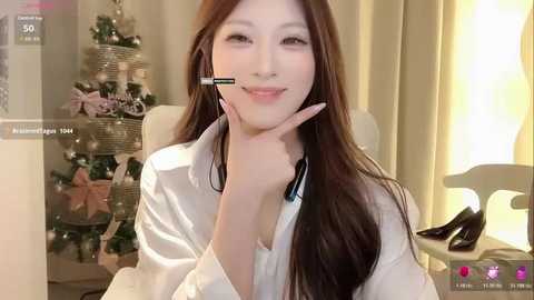 Video of an Asian woman with long brown hair, wearing a white shirt, headset, and makeup, seated in a cozy room with a Christmas tree and lamp.