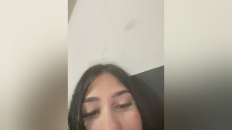 A video showing a close-up of an Asian woman with dark hair, wearing makeup, and looking downward. The background is a plain, off-white wall with a small, circular stain. The image is slightly blurred.