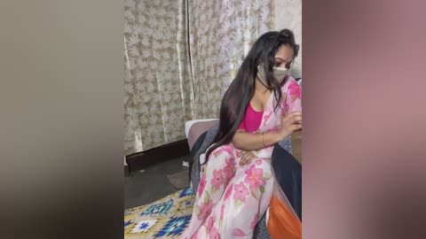 Media: Video of a woman in a pink floral sari with a face mask, seated on a couch, surrounded by patterned curtains and a colorful rug.