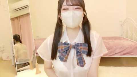 Video of a young Asian woman with long black hair, wearing a white blouse with a plaid bow, face mask, and seated on a bed in a simple, beige room with a mirror.