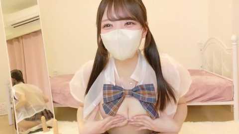 Video of an East Asian woman with long black hair and a white face mask, wearing a white blouse with a blue plaid bow, standing in a cozy, pink-themed bedroom.