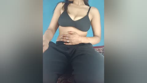 Media: A video of a light-skinned woman with long dark hair, wearing a black bra and pants, sitting on a patterned chair with her hand on her belly.