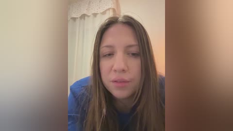 Media: Video of a young Caucasian woman with light skin and long, straight brown hair, wearing a blue shirt, looking slightly sad, indoors with a beige curtain and a lampshade in the background.