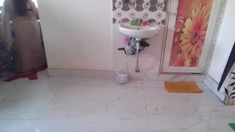 Video of a small, cluttered bathroom with a white ceramic sink against a floral-patterned wall, a beige curtain, and a yellow towel on the floor.