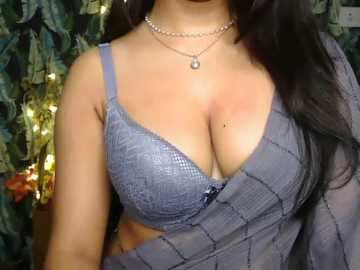 Video of a woman with long dark hair, wearing a light blue lace bra and a sheer gray shawl, adorned with a pearl necklace. Background shows a dark, patterned curtain and a string of fairy lights.