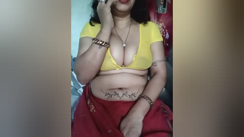 A video of a South Asian woman with light skin, wearing a yellow bra, red skirt, and bracelets, sitting in a red chair, showing a tattoo on her abdomen.