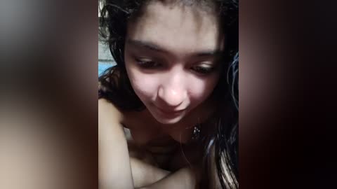 Video of a young woman with wet, dark hair and light skin, smiling slightly, wearing a necklace, in a dimly lit room.