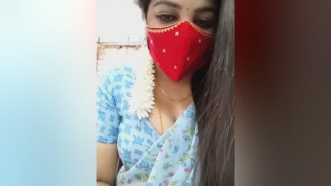 A video of a woman with long black hair, wearing a red mask, a floral-patterned blue saree, and a white flower necklace, standing in front of a white wall with a wooden hanger.