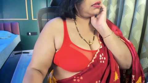 Media: Video of a South Asian woman with medium skin tone, wearing a red sari with white floral patterns, a matching red bra, and a gold necklace. She sits in a modern bedroom with a green wall, a white bed, and patterned curtains.