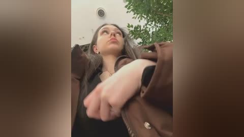 Video of a woman with straight, dark hair, wearing glasses and a brown jacket, looking up with a slight frown, captured from a low angle.