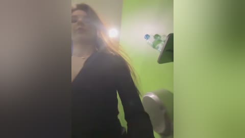 Video of a young woman with long brown hair, wearing a black jacket, standing in a dimly lit bathroom with green walls, a white soap dispenser, and a green towel rack.