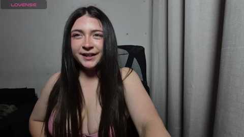 A video of a young Caucasian woman with long, straight brown hair, wearing a pink bra, smiling, sitting in a chair with beige curtains in the background.