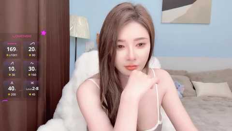 A video of a young Asian woman with long, straight brown hair, sitting on a bed in a cozy bedroom. She wears a white camisole and looks contemplative. The room has a soft, pastel color scheme.