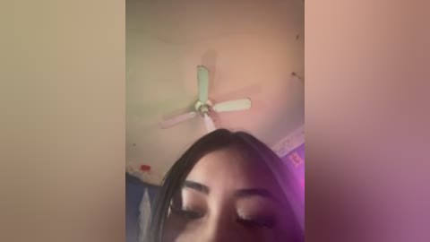 A video of a woman with long, dark hair, partially cropped out, looking upwards. A ceiling fan with light-colored blades is visible above. The background includes a wall and a blurred object.