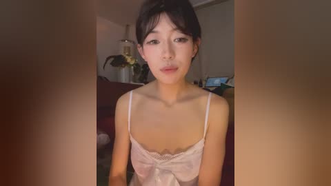 Video of an Asian woman with short black hair, wearing a white lace camisole, standing indoors with blurred background.