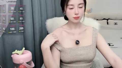 Video of an Asian woman with fair skin, dark hair in a bun, wearing a beige ribbed top, with a pink purse and a plush toy on a grey chair.