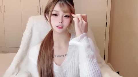 A video of a young Asian woman with long, straight brown hair in pigtails, wearing a white knitted sweater, sitting on a white bed, adjusting her hair. Background includes beige wardrobe doors.