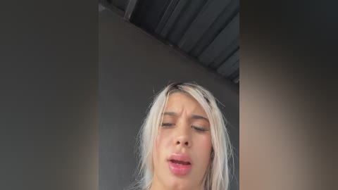 Video of a young woman with platinum blonde hair, closed eyes, and slightly open mouth, possibly in pain or discomfort, against a plain gray wall and a metallic ceiling.