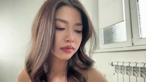 Video of a young East Asian woman with long, wavy brown hair, fair skin, and soft features. She wears a pink lipstick and looks down, in a bathroom with a white window and radiator.