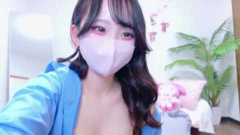 Video of an Asian woman with long dark hair, wearing a blue shirt and white mask, standing in a room with a plant and plush toys.