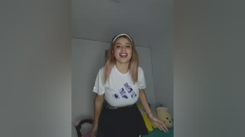 Video of a young, smiling Asian woman with long, straight, pink hair, wearing a white T-shirt with blue geometric patterns, black skirt, and white headband, standing in a dimly lit room with white walls, a stuffed toy, and a yellow object in the background.