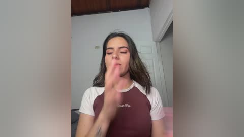 Video of a young woman with long, dark hair, wearing a maroon and white athletic top, licking her finger, in a dimly lit room with white walls and a wooden ceiling.