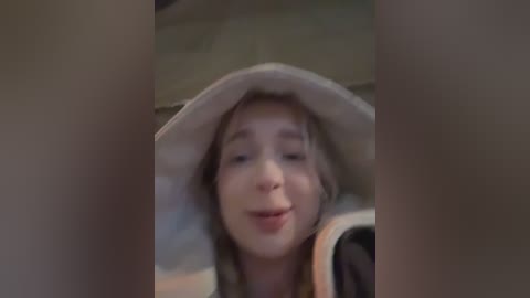 A blurry video of a young girl with blonde hair wearing a light-colored hat, smiling, partially obscured by out-of-focus hands.