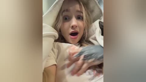 A video of a shocked young woman with light skin and blonde hair, wearing a beige t-shirt, reacting to a bloody, severed hand.