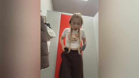 A video of a young Asian woman with blonde braided hair, wearing a white graphic T-shirt and high-waisted brown pants, adjusting her clothes in a brightly lit, white-tiled bathroom with red accents.