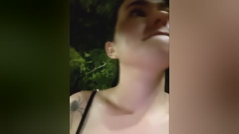 Video of a young woman with short dark hair and a black tattoo on her right shoulder, gazing upwards, partially obscured by dark foliage.